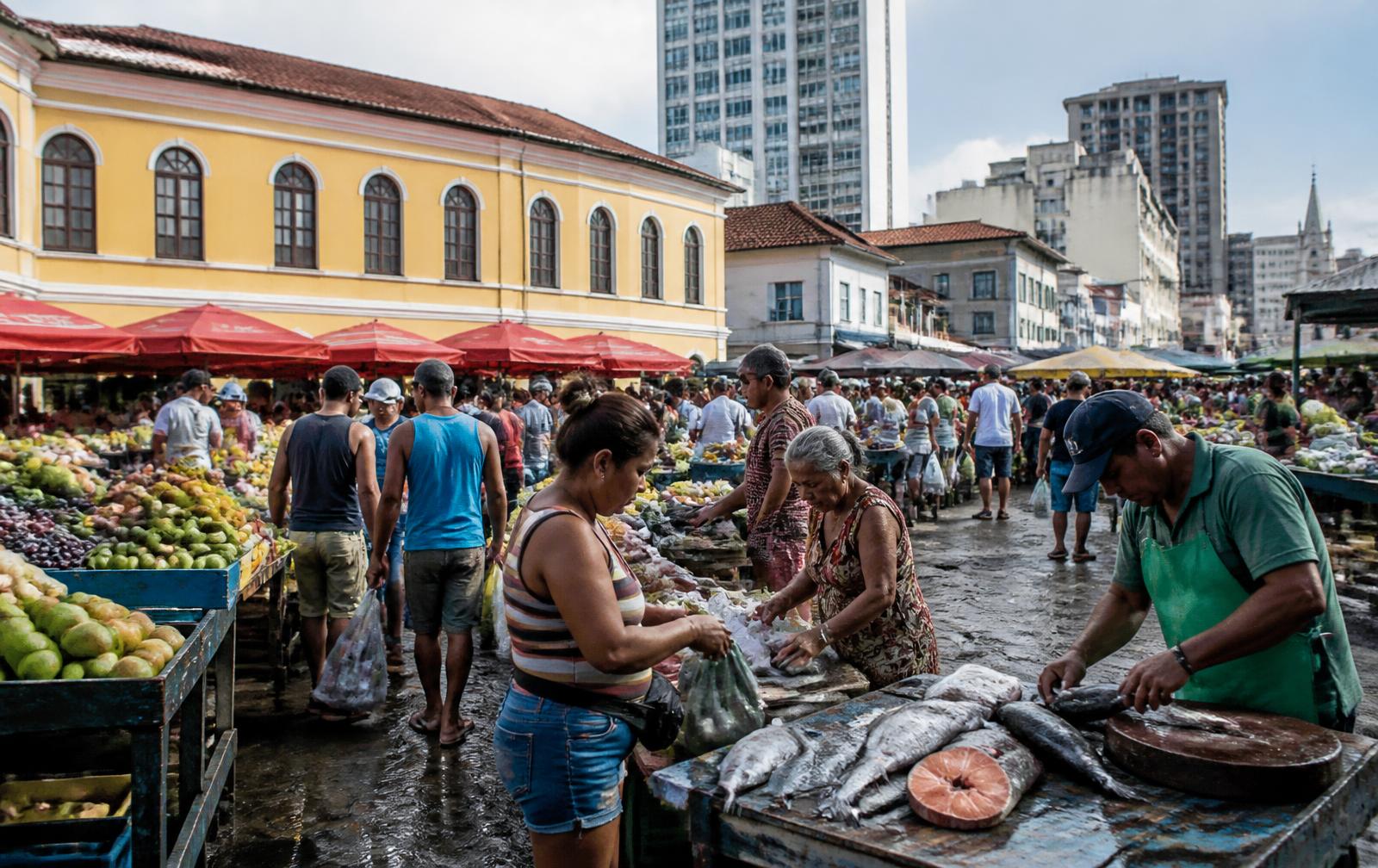 Feira urbana movimentada em Belém, com bancas de frutas e peixes, prédios e arquitetura histórica ao fundo