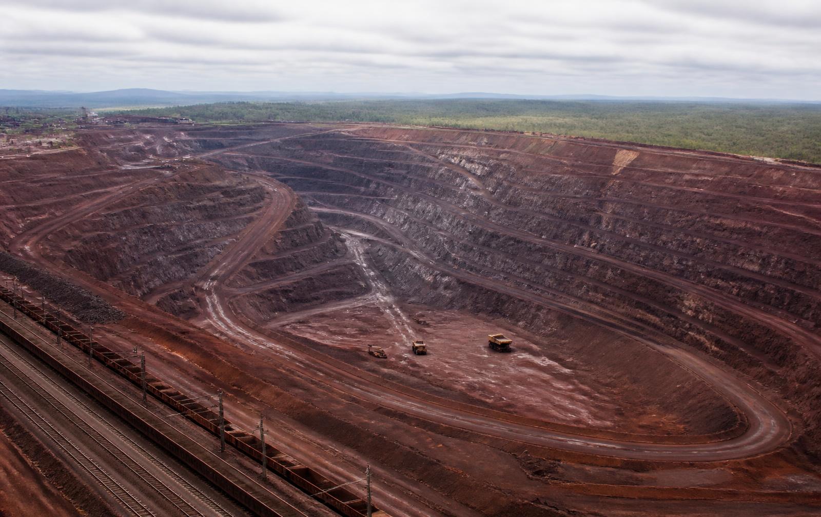 Grande mina a céu aberto com bancadas de terra avermelhada, caminhões e uma ferrovia de carga na borda